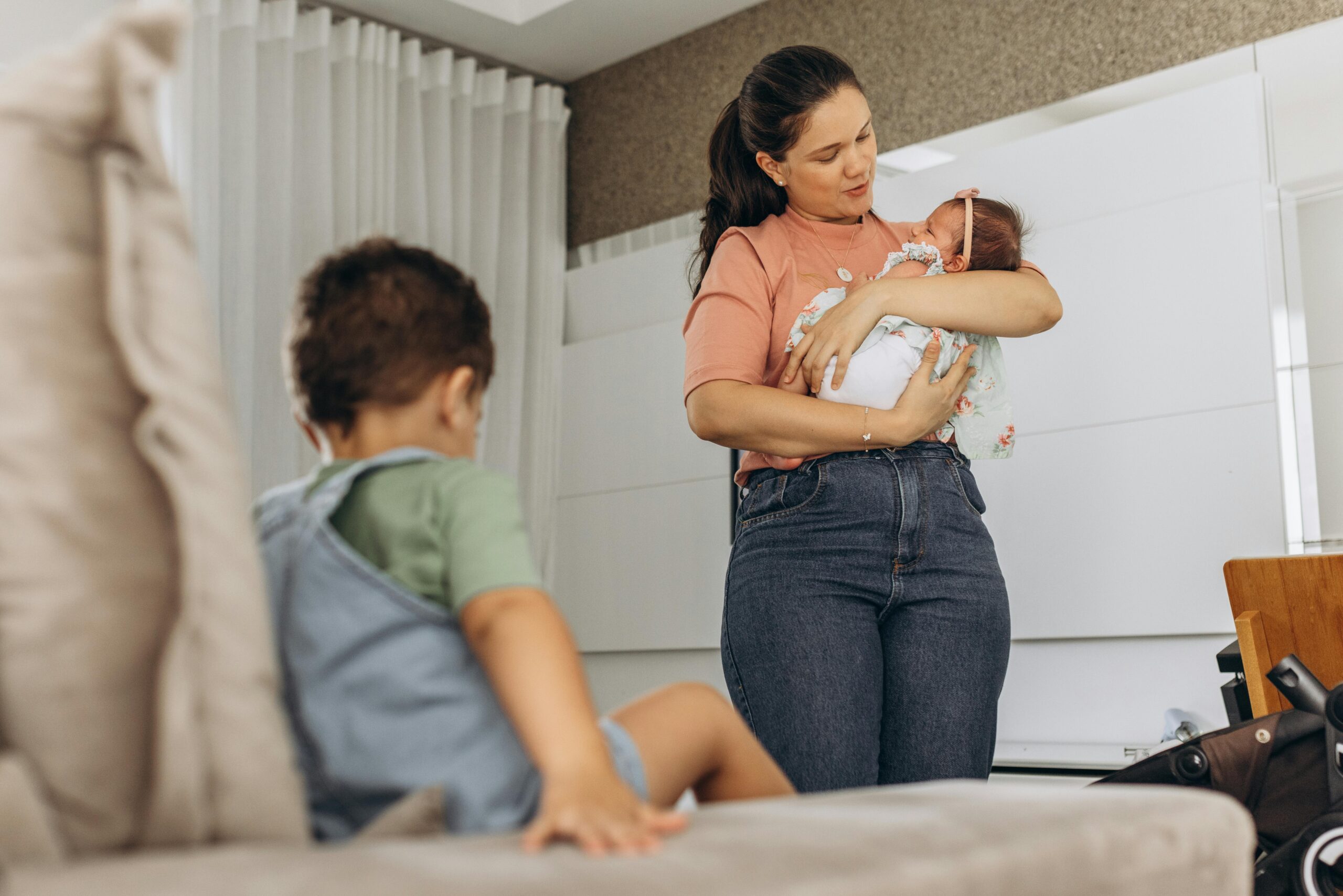 A mother lovingly cradles her baby while her child sits nearby on the couch in a modern room setting.